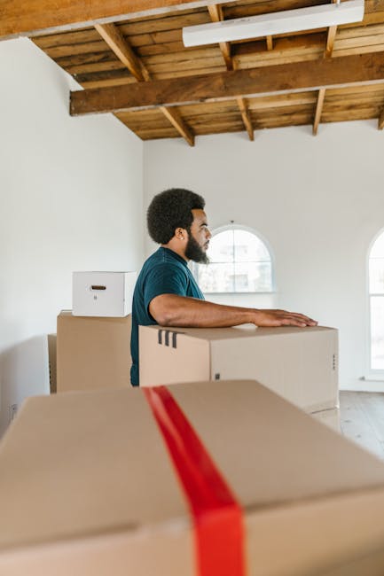 A man with dark curly hair and a beard is standing inside a bright, unfurnished room with large arched windows and light-colored wooden flooring, engaged in house relocation activities. He is wearing