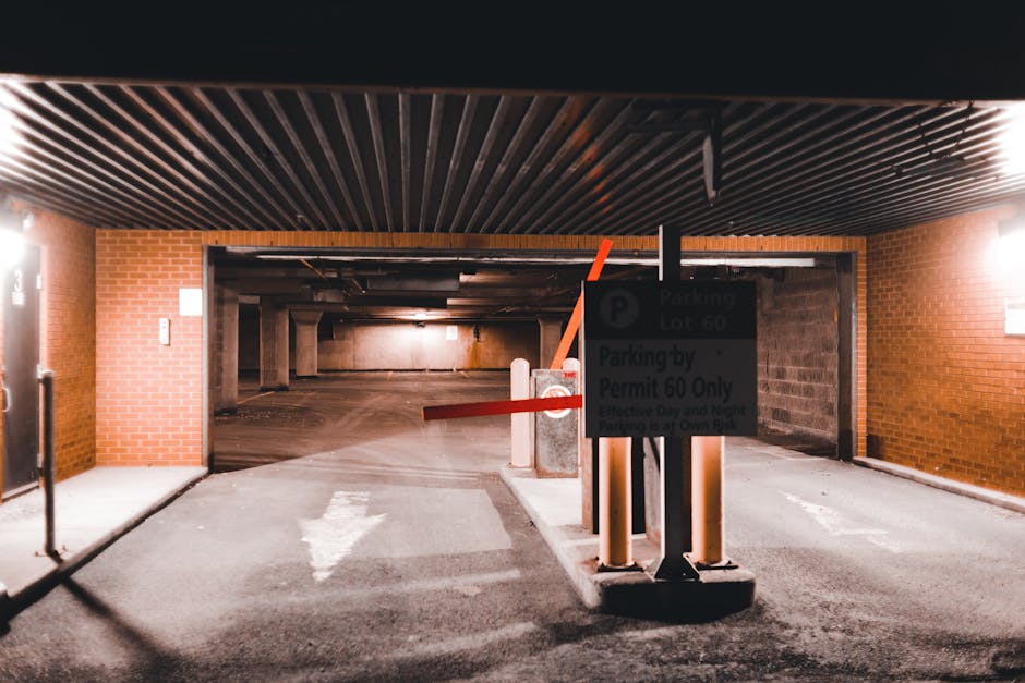The image shows an indoor parking garage entrance with a controlled access barrier and a parking permit sign attached to a black post. The barrier is red and white, and is in a raised position, allowing vehicles to pass through, with a white arrow painted on the concrete ground indicating the direction for vehicle movement. The parking permit sign displays parking lot information and restrictions, including 'Parking Lot 60' and 'Parking by Permit 60 Only,' with some text slightly obscured by lighting. The garage walls are made of red brick on either side of the entrance, and the ceiling features wooden slats with installed lighting fixtures casting illumination inside the space. Inside, the parking area extends further, with concrete pillars and colored lighting creating a dimly lit environment suitable for vehicle storage and home relocation logistics. The surrounding area appears clean and organized, reflecting a secure site often used during packing, loading, and furniture transport processes conducted by professional removals services such as Man and Van Kilburn to facilitate house moves in the Kilburn area.