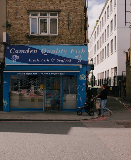 The image shows the storefront of Camden Quality Fish, a seafood shop with a prominent blue sign displaying the business name and images of fish. The shop has a large glass display window showcasing various fresh and frozen seafood products inside, with some packaging and ice visible. Above the entrance, there's a smaller sign indicating the shop offers fresh fish, sea food, and original food items. To the right of the shop, a person dressed in black is pushing a baby stroller on the sidewalk, carrying a bag. They are positioned adjacent to a narrow street or alleyway with modern white residential or commercial buildings lining the background. The scene is outdoors during daylight, and the setting appears to relate to household or retail relocation activities, aligning with house removals and moving services, with the shop potentially being part of the logistical environment for a move or delivery scheduled in Kilburn, as referenced in the Camden Council moving day rules page. Man and Van Kilburn occasionally provides assistance with furniture transport, packing, and loading process in such environments.