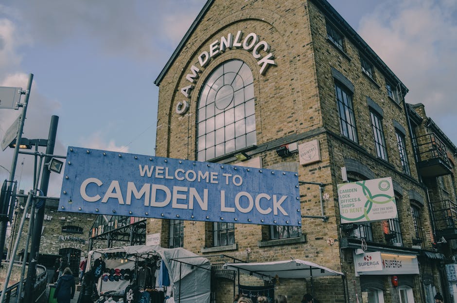 Exterior view of the Camden Lock building featuring a large, white, arched sign with the words 'CAMDEN LOCK' mounted on the brick facade under a cloudy sky. A sizeable blue banner reading 'WELCOME TO CAMDEN LOCK' is positioned in front of the building, attached to black metal fencing. The scene includes a small outdoor market stall covered with a white canopy, displaying clothing and accessories beneath, with some customers nearby. To the right, there is a green and white sign advertising Camden Lock offices and shops available for rent, mounted on the building's wall. Visible are several black metal fire escapes and balconies along the building, and additional signage indicating the location as part of Camden's market and shopping district. The environment suggests a busy, well-lit area with natural daylight, appropriate for a house or furniture move, with visible preparation for loading or transport activities as part of home relocation or furniture transport services, consistent with house removals in Kilburn.