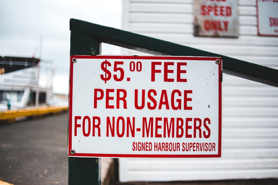 Close-up view of a white rectangular sign with bold red lettering attached to a dark green post at a harbour or marina. The sign states a $5.00 fee per usage for non-members, signed by the harbour supervisor. In the background, there are partially visible boats docked at the harbour with white hulls, moored alongside a paved area. The image suggests a setting related to boat or vessel access, which could involve moving or loading equipment as part of a home relocation or transport service. The environment appears overcast with diffused natural light, and the focus is primarily on the sign and the surrounding dock area, relevant to transport logistics and maritime loading processes occasionally associated with removals or furniture transport in a house moving context.