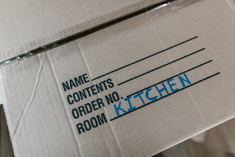 Close-up view of a cardboard moving box with printed labels for a home relocation. The box has sections labeled 'Name,' 'Contents,' 'Order No.,' and 'Room,' with 'Kitchen' handwritten in blue marker in the 'Room' section. The box is partially sealed with clear packing tape and appears to be placed on a surface inside a property, likely near an entryway or in a corridor. The packaging materials include standard brown cardboard and plastic tape, and the box is used for packing kitchen items during a furniture transport or packing and moving process. The environment has neutral lighting, and the image emphasizes organized packing, typical in house removals managed by services like Man and Van Kilburn.