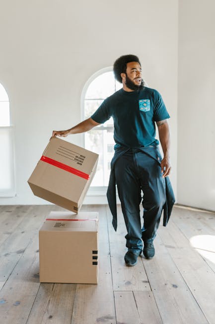 A man with dark skin and a beard is inside a bright, empty room with white walls and a large arched window that allows natural light to fill the space. He is wearing a blue uniform with a logo on the chest, indicating his role in house removals or furniture transport services. The man is holding a cardboard box with red packing tape on top, preparing to load it onto another box placed on the wooden floor. Nearby, there are additional cardboard boxes, some stacked, which are likely packed for home relocation. The room's minimal furnishings and natural lighting suggest a house or apartment being prepared for moving day. The scene captures the process of packing and loading items as part of a professional moving service, reflecting the logistics involved in packing and furniture transport during house removal projects.