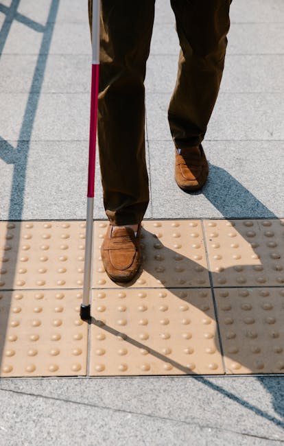 Close-up view of a person standing on a tactile paving surface outside a building, with brown leather shoes and dark trousers visible. The individual is holding a white cane with a pink tip, indicating they may have a visual impairment. The tactile paving consists of beige tiles with raised bumps, set on a grey pavement, and is designed to assist with navigation. Shadows cast by the person and cane are visible on the ground. In the background, part of a doorway or entrance area is seen, with a glimpse of the interior flooring. This scene reflects elements of accessible movement and orientation, relevant to home relocation or moving services such as those offered by Man and Van Kilburn, where safe and accessible passage during packing and furniture transport is essential.