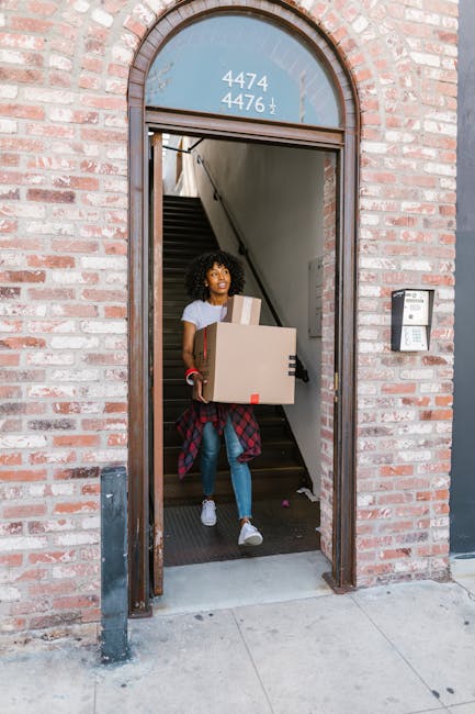 A woman with curly dark hair, dressed in a white t-shirt, blue jeans, and white sneakers, is seen carrying a cardboard box with red and black tape out of a brick building entrance. She is stepping down from the doorway onto a paved sidewalk during a home relocation or furniture transport process. Inside the building, a staircase with black treads and white risers leads upward, and a small section of a handrail is visible along the wall. The building features a curved brick arch framing the entrance, with a glass panel displaying the numbers 4474, 4476, and a vertical arrow. To the right of the doorway, a black intercom or access control panel is mounted on the brick wall. The scene is well-lit, indicating daytime, and the surroundings suggest a moving or removals service of a residential property, consistent with professional house relocation activities, with the company Man and Van Kilburn possibly involved.