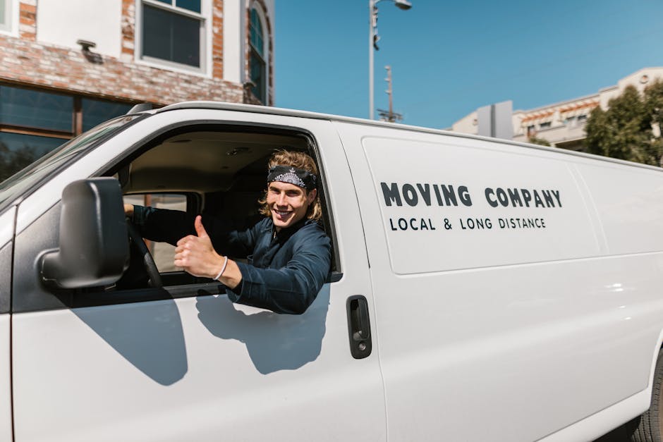 Two men involved in a home relocation process are loading a cardboard box into a white van with open rear doors, parked on a paved driveway next to a brick wall. One man is wearing a dark baseball cap