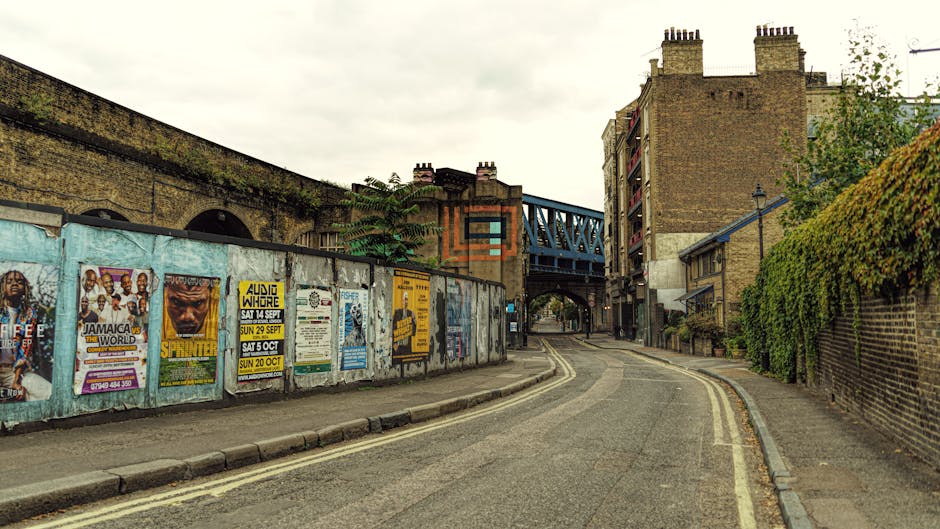 A narrow residential street in Kilburn with a gently curving asphalt road marked by double yellow lines along the edge. On the left side, a weathered metal fence covered in colorful posters and advertisements runs parallel to the pavement, separating the street from an elevated railway bridge painted in dark blue with a metal framework. Underneath the bridge, there is an arched passageway. To the right, a brick wall topped with lush green ivy borders the sidewalk, which is lined with a row of small, multi-story brick houses featuring chimneys and balconies. These residential buildings vary in height and style, with some having metal railings and window shutters. The scene appears to be captured during daylight hours with overcast skies, and there are no visible pedestrians or vehicles. The setting suggests a quiet, urban neighbourhood suitable for house removals and furniture transport, with potential for loading and unloading activities related to home relocation by local removals services such as Man and Van Kilburn.