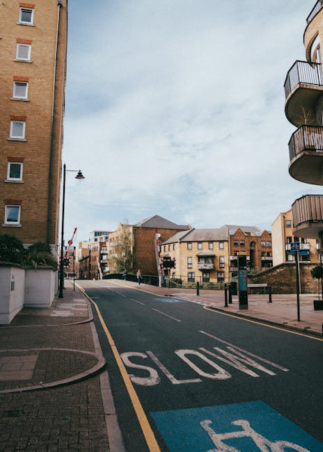 A residential street scene captured during the house removal process, featuring a paved road with a visible 'SLOW' sign painted on the asphalt, and a designated parking bay marked with blue paint. On the left side of the image, there is a multi-storey brick building with several windows, while on the right, a modern apartment with curved balconies is partially visible. In the background, beyond the street, there are additional buildings with flat roofs and varying architectural styles, under a sky filled with diffuse clouds. A man, dressed in casual clothing, is seen walking along the sidewalk near the buildings. The scene suggests an urban environment suited for home relocations, with the street cleared for vehicle access and parking, and no active loading or moving activity visible at the moment. Man and Van Kilburn occasionally works in such street environments to facilitate efficient furniture transport and packing and moving services for flats along Kilburn High Road.