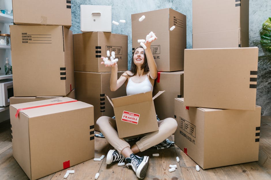 A young woman sitting on a wooden floor surrounded by numerous cardboard moving boxes of various sizes, some sealed with red and black tape, inside a room with a textured light blue wall. She is smiling and tossing packing foam peanuts into the air, with a partially opened box on her lap revealing more packing material. In the background, a small green plant is visible on the right, and part of a storage shelf or kitchen counter with a water bottle is seen on the left. The scene depicts a packing process typical of home relocation or furniture transport, with items ready for moving. The environment is well-lit, and the arrangement suggests an active packing or loading phase, with [COMPANY_NAME] providing professional removals services as indicated by the context of the image.