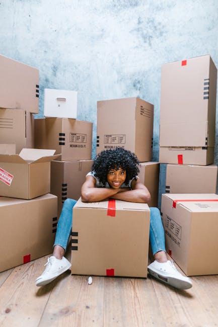 A young woman with curly black hair, dressed in a white top and light blue jeans, is sitting on the wooden floor of an interior space surrounded by numerous cardboard boxes of various sizes, some sealed with red tape and others partially open. She is resting her arms on a large moving box and smiling at the camera. The background features a plain, textured light blue wall, and the boxes are stacked against it, indicating an ongoing home relocation or packing process. The scene depicts the packing stage of furniture transport and house removals, with the woman in a relaxed pose amidst moving supplies, supporting the context of professional removals services such as those provided by Man and Van Kilburn.