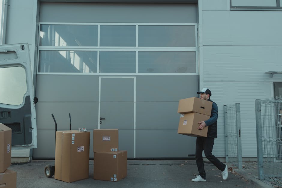 A man wearing a cap, dark jacket, and trousers is outside a commercial building, carrying two cardboard boxes stacked in his arms during a furniture transport process. The building has large grey metal panel walls and a large windowed garage door. On the pavement in front of the building, several cardboard boxes of various sizes are placed, some upright and others on their sides, with labels and symbols indicating fragile contents. A hand truck or dolly with two handles is positioned next to a smaller box, suggesting recent or ongoing loading or unloading. To the left, part of a white van with an open rear door is visible, likely used for home or business relocation services. The scene is set in daylight, with natural light illuminating the area, demonstrating a typical moving or packing operation as part of a professional removals service by Man and Van Kilburn, supporting house removals or commercial moves.