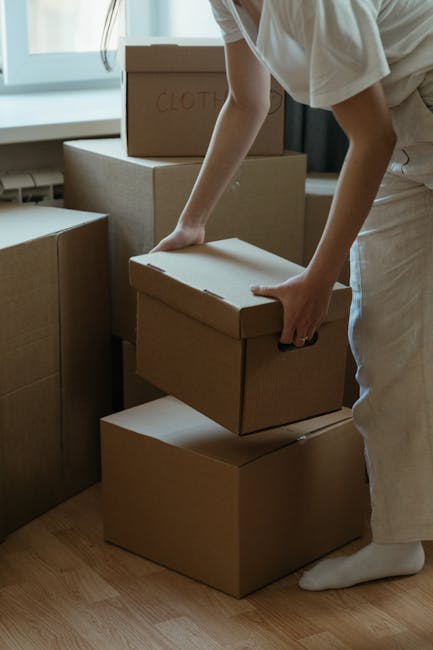 A person wearing light-colored clothing is seen in an indoor space, carefully lifting a small cardboard moving box with both hands, positioning it on top of a larger box, amidst a stack of other cardboard boxes. Some boxes have handwritten labels such as 'CLOTHES.' The scene suggests active packing or unpacking as part of a home relocation process. The area is well-lit with natural light from nearby windows, and the boxes are placed on a wooden floor. This image reflects furniture transport, packing and moving activities, and the logistics involved in house removals, illustrating the services offered by Man and Van Kilburn for small business office removals or residential moves.