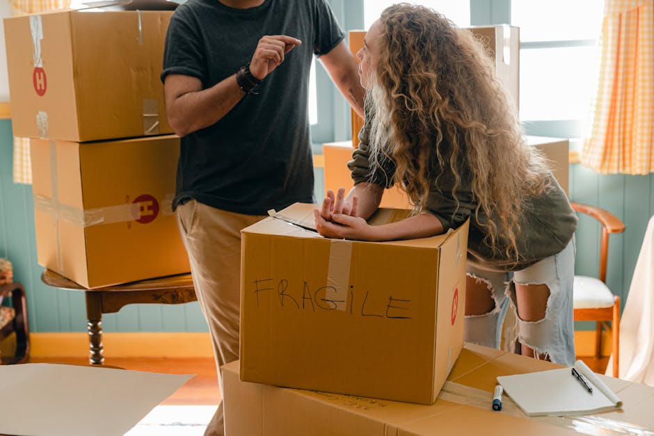 A person with a dark t-shirt and khaki pants is standing inside a room filled with cardboard moving boxes, some stacked on a wooden table and others placed on the floor, with additional boxes visible in the background. A woman with curly blonde hair, wearing a dark long-sleeved top and ripped white jeans, is leaning over a medium-sized cardboard box labeled 'FRAGILE' with handwritten black marker, preparing for a home relocation. The scene shows packing and moving activities, with the woman holding the box's top flaps open while the person appears to be giving instructions or guidance. The room is illuminated by natural light coming through windows with orange checkered curtains, and there are plastic wrap and packing materials nearby, indicating an ongoing packing process as part of furniture transport or a house move.