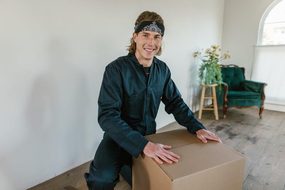 A person with a dark t-shirt and khaki pants is standing inside a room filled with cardboard moving boxes, some stacked on a wooden table and others placed on the floor, with additional boxes visible in the background. A woman with curly blonde hair, wearing a dark long-sleeved top and ripped white jeans, is leaning over a medium-sized cardboard box labeled 'FRAGILE' with handwritten black marker, preparing for a home relocation. The scene shows packing and moving activities, with the woman holding the box's top flaps open while the person appears to be giving instructions or guidance. The room is illuminated by natural light coming through windows with orange checkered curtains, and there are plastic wrap and packing materials nearby, indicating an ongoing packing process as part of furniture transport or a house move.
